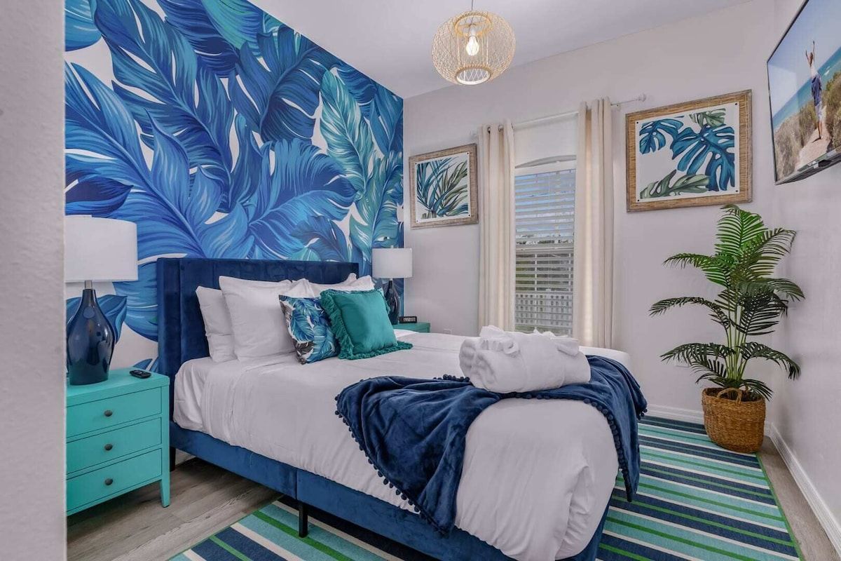 Een slaapkamer met een grote muurschildering van een tropisch blad, een bed met witte lakens, blauwgroene kussens en een gestreept vloerkleed.