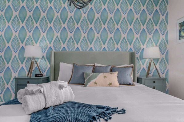 A neatly made bed with a blue blanket and pillows, two bedside tables with lamps, and a patterned wallpaper.