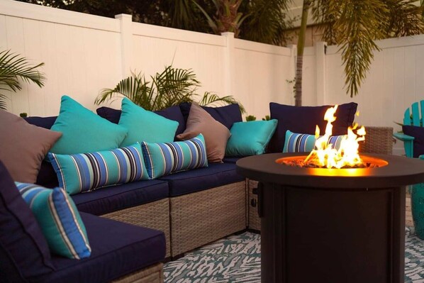 An outdoor seating area with a fire pit, cushioned seating, and a white fence.