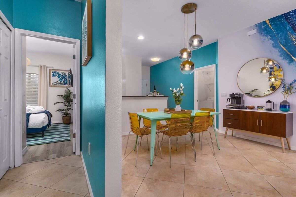 A dining area with a table and chairs, a kitchen in the background, and a bedroom with a bed and window.