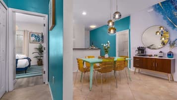 A dining area with a table and chairs, a kitchen in the background, and a bedroom with a bed and window.
