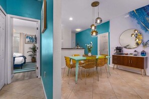 A dining area with a table and chairs, a kitchen in the background, and a bedroom with a bed and window.