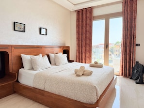 A hotel room with a large bed, wooden headboard, two framed pictures, a balcony with curtains, and a standing fan. - Stylish Apt Steps From the Beach (Essaouira)