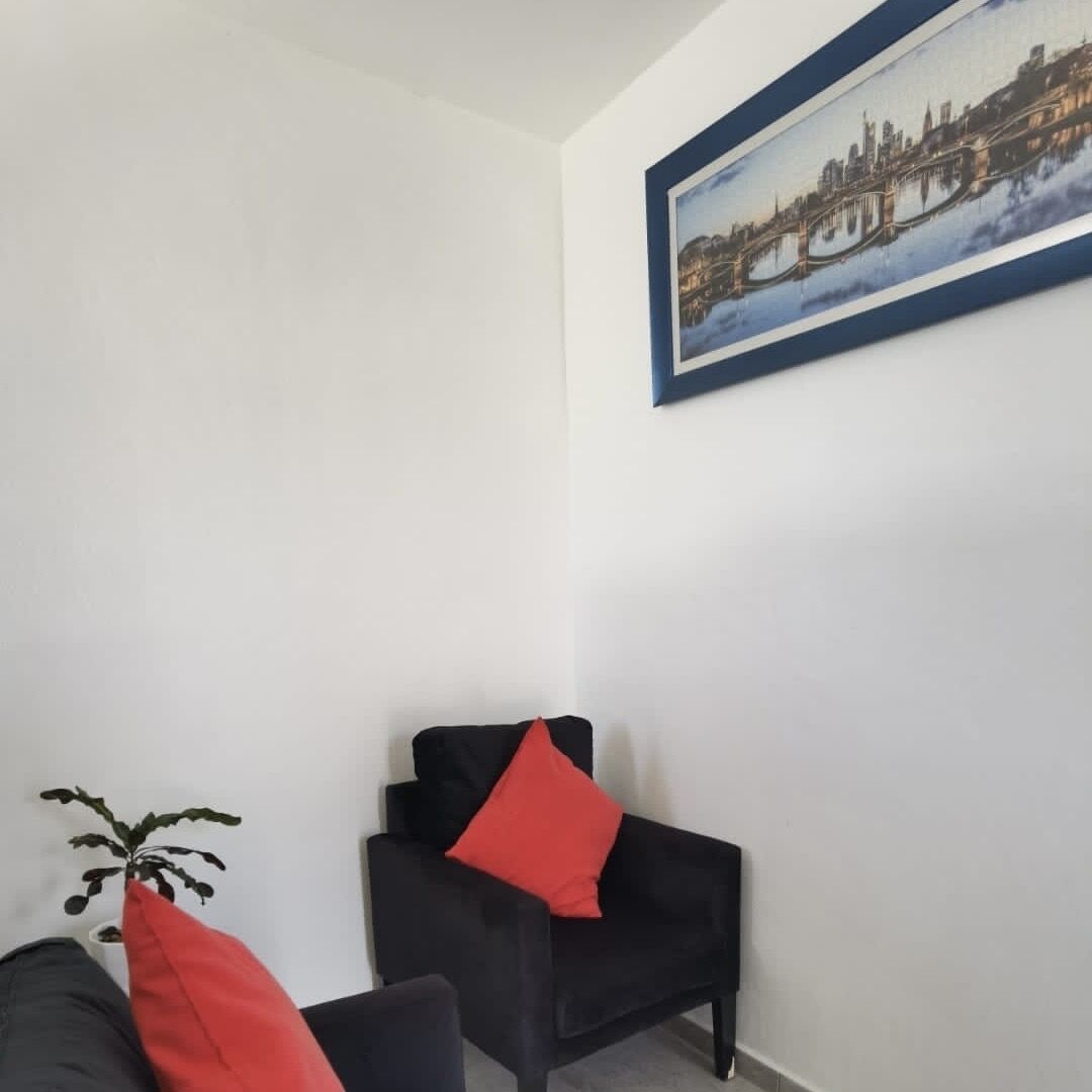 A room with a black armchair, a small table with a plant, and a framed picture of a cityscape on the wall.