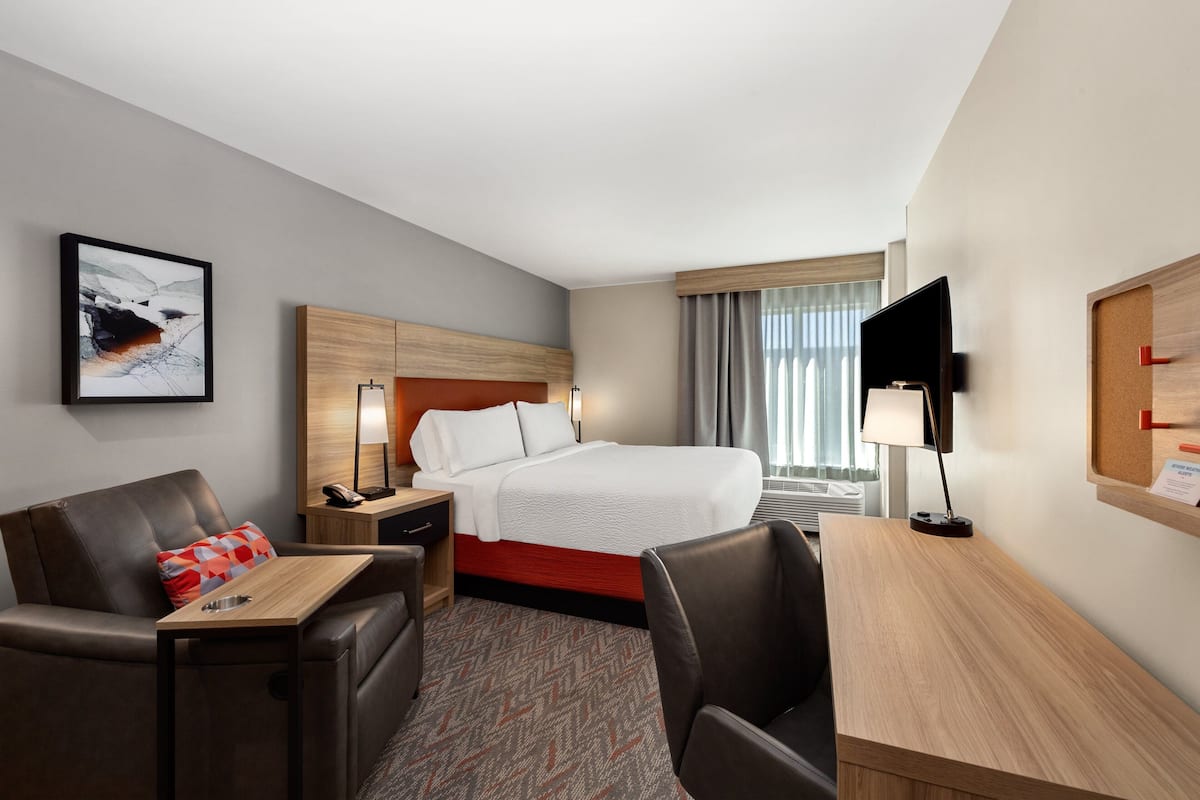 A hotel room with a bed, a desk, a chair, a nightstand, a lamp, a television, and a framed picture on the wall.