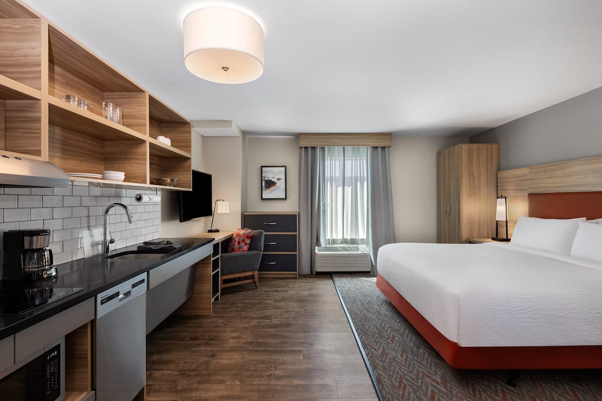 A modern hotel room with a kitchen area, a bed, a desk, and a window with curtains.