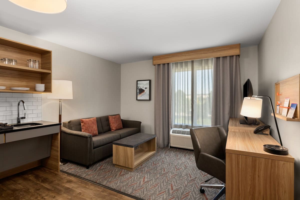 A compact hotel room with a kitchenette, a sofa, a desk, and a large window with curtains.