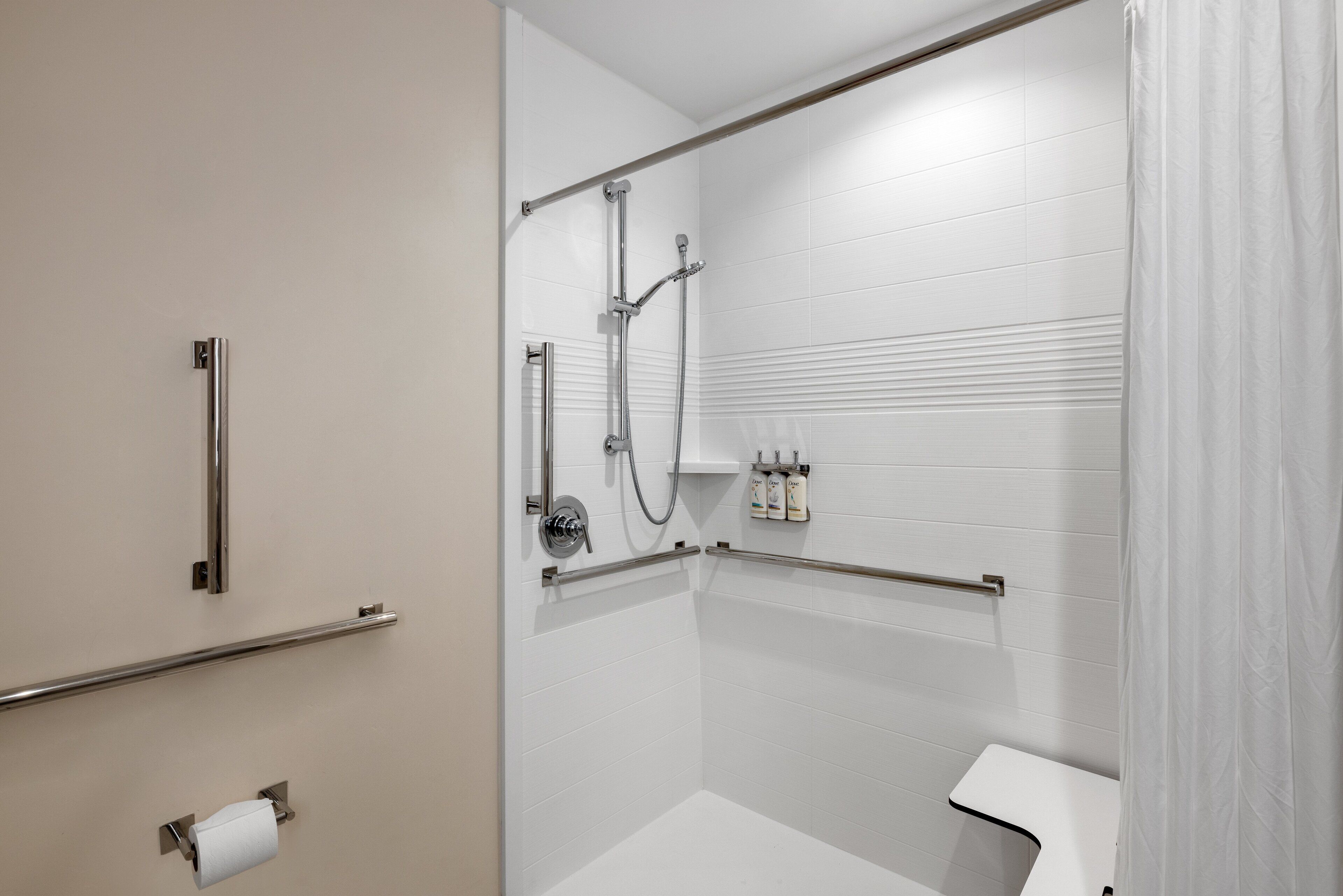 Un baño moderno con paredes de azulejos blancos, una ducha con cabezal de ducha de mano, inodoro y barandilla.