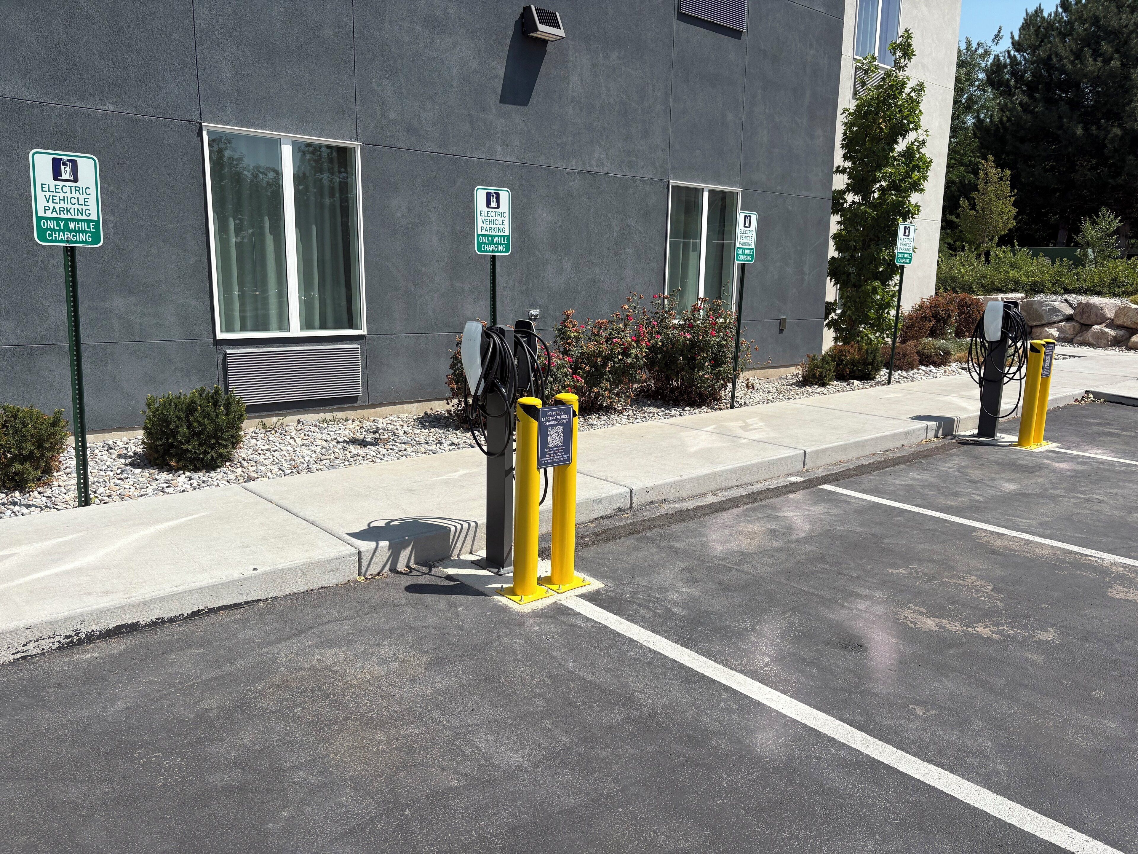 Estaciones de carga para vehículos eléctricos con señalización de estacionamiento, un edificio con ventanas amplias y una zona de estacionamiento pavimentada.