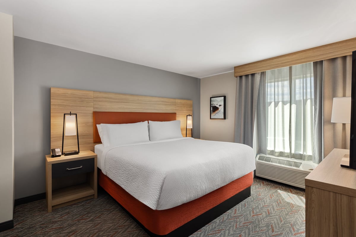 A hotel room with a large bed, a nightstand, a desk, a chair, a window with curtains, and a wall-mounted artwork.