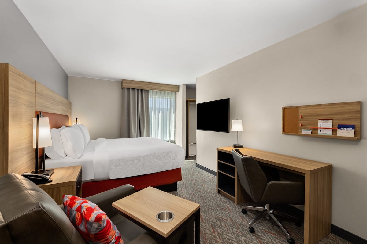 A hotel room with a bed, a desk, a chair, a TV, and a bulletin board.