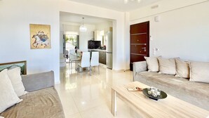 A modern living room with a sofa, a coffee table, and a dining area with chairs. There is a kitchen visible in the background. - Luxury Villa San George with private pool (Perigiali)