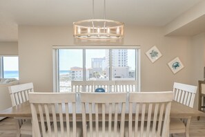 Ein Essbereich mit einem Holztisch und Stühlen, einer Hängeleuchte und Blick auf den Strand und Gebäude durch große Fenster.