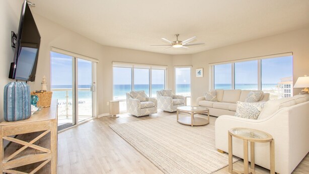 Ein geräumiges Wohnzimmer mit großem Fenster zum Strand, Deckenventilator und flachbildschirm-Fernseher an der Wand.
