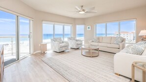 Ein geräumiges Wohnzimmer mit einem großen Schiebeglasfenster, das auf einen Balkon führt, einem Deckenventilator und Blick auf den Strand.