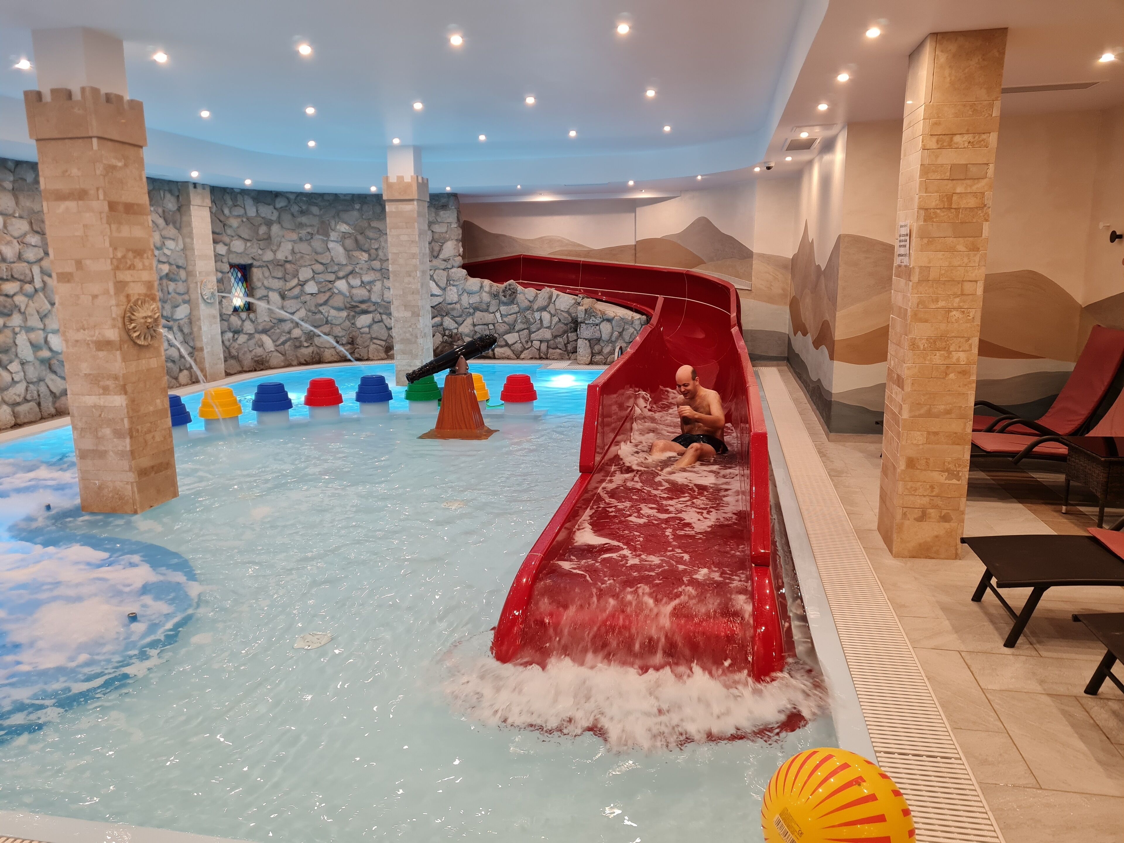 Un tobogán acuático interior con un resbaladizo de color rojo y una zona de piscina con una pared de piedra y un mural.