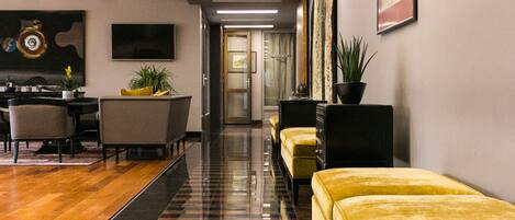 A modern hotel lobby with a long reception desk, several yellow armchairs, a dining area with a table and chairs, and a large painting on the wall.