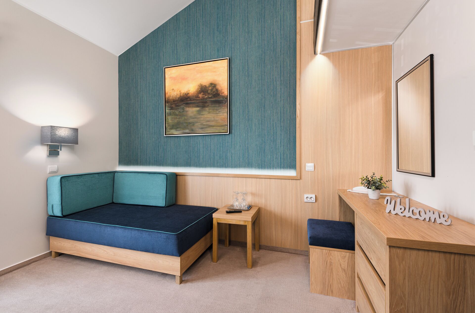 A modern room with a teal accent wall, a bed with a blue headboard, a small wooden table with a vase, and a desk with a 'Welcome' sign.