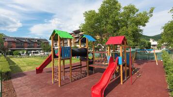 A playground with slides and climbing structures, surrounded by a hotel and greenery.