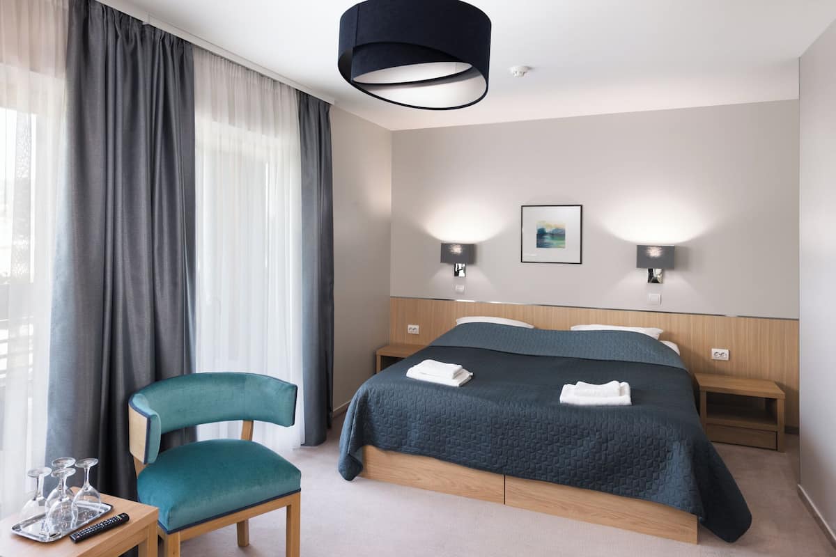 Hotelkamer met een bed, twee nachtkastjes, een stoel, een bureau met dienblad en afstandsbediening, en een groot raam met gordijnen.