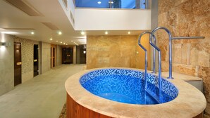 Una piscina interior moderna con borde de madera y fondo de mosaico azul, rodeada de paredes de piedra y un techo alto con iluminación empotrada.