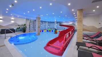 Una piscina interior con tobogán, sillas de descanso y un muro de piedra de fondo.