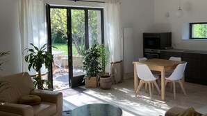 Una sala de estar moderna con un sofá, una mesa de centro redonda y un comedor con sillas y una mesa. Hay una zona de cocina con estufa y refrigerador. Amplias ventanas dejan entrar la luz natural.