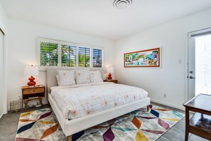 Quarto com cama, duas mesas de cabeceira, janela com persianas, tapete colorido e um quadro pendurado na parede.
