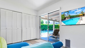 Una habitación con un amplio ventanal corredero que da a una zona de piscina, un televisor de pantalla plana en la pared y un sofá azul con cojines estampados.