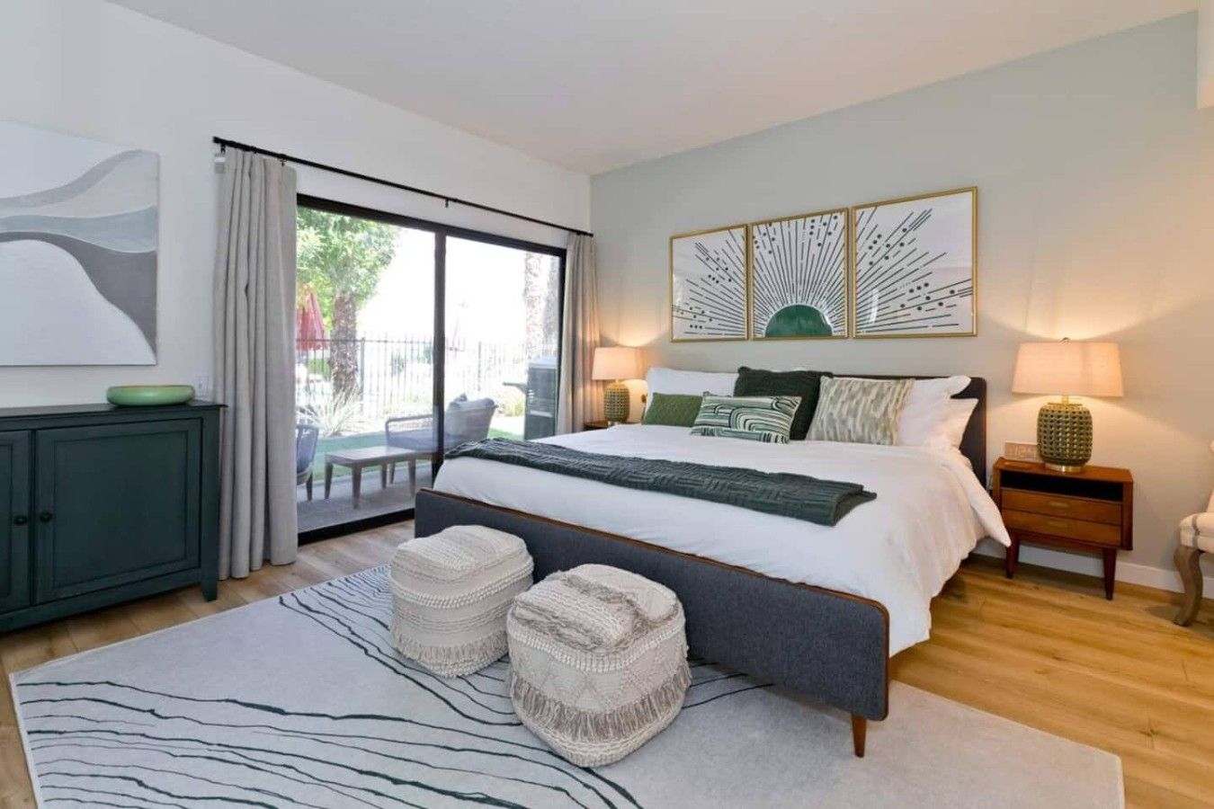 A modern bedroom with a large bed, a sliding glass door to a balcony, and a view of greenery outside.