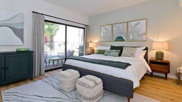 A modern bedroom with a large bed, a sliding glass door to a balcony, and a view of greenery outside.
