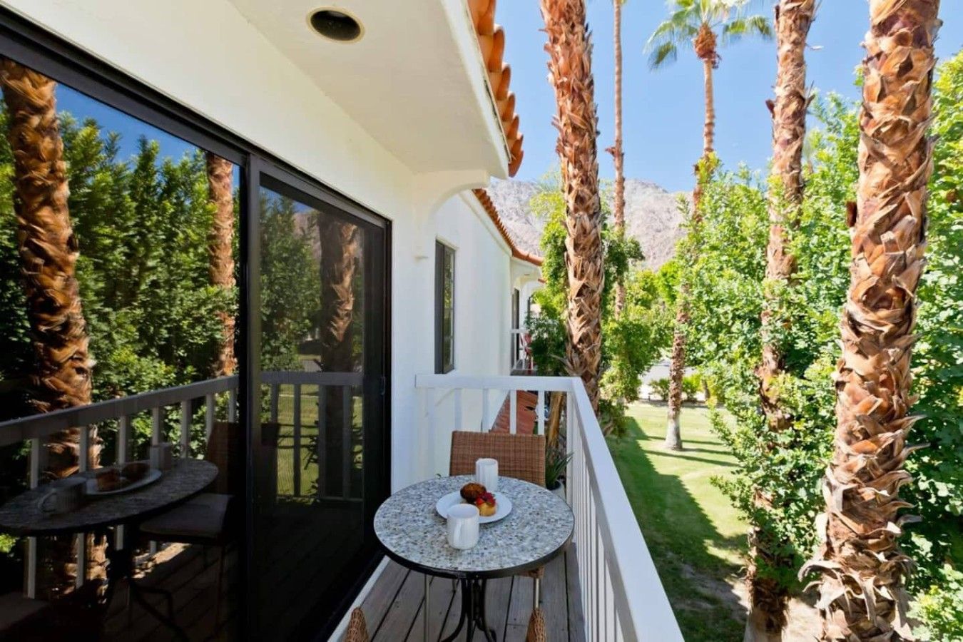 A balcony with a table and chairs, overlooking a garden with palm trees and a mountain in the background.