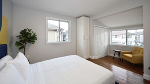 A bedroom with a bed, a plant, a yellow armchair, and a view of the city through large windows. - Luke 2 House, Stylish, Boston, Parking, Sleeps 8, Memorable  (Everett)