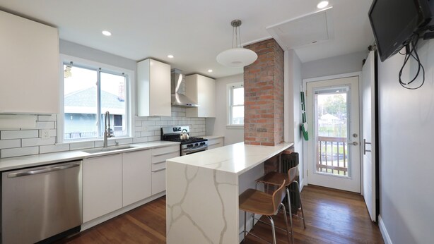 A modern kitchen with white cabinetry, a marble island, and a brick accent wall. - Luke 2 House, Stylish, Boston, Parking, Sleeps 8, Memorable  (Everett)