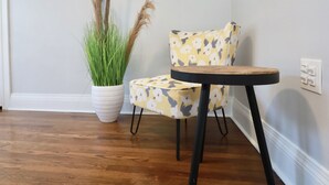 A corner room with a wooden floor, a white potted plant with green grass, a yellow floral chair, and a small round table with black legs. - Luke 2 House, Stylish, Boston, Parking, Sleeps 8, Memorable  (Everett)