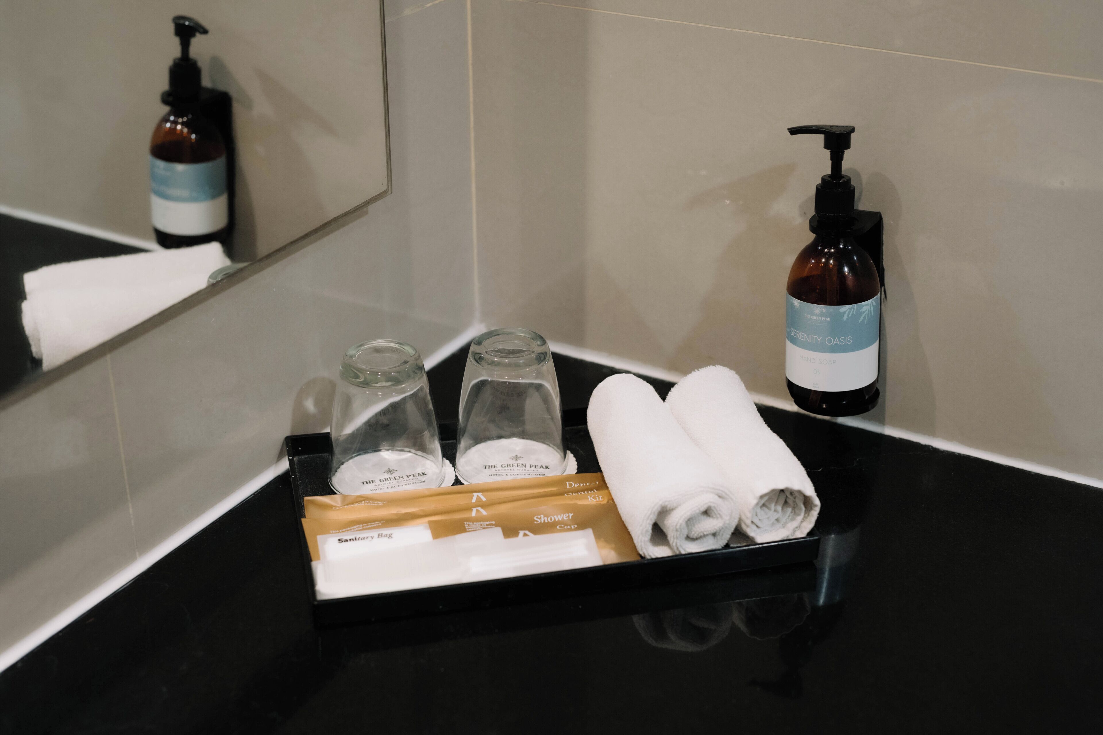 A bathroom with a black countertop, a towel rack, a mirror, and toiletries including a bottle of shampoo, a bottle of conditioner, and a shower kit.