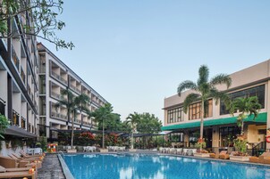 Una piscina con sillas de descanso y un área de estar cubierta, rodeada de edificios con balcones y palmeras.