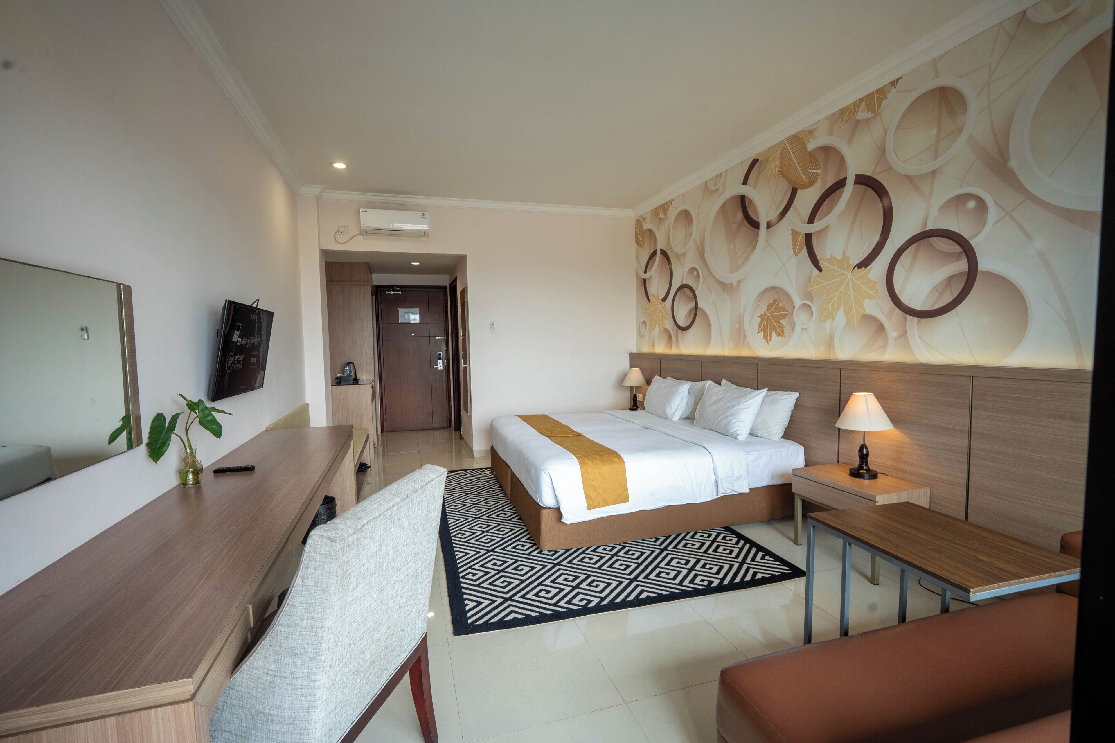A hotel room with a bed, bedside table, lamp, desk, chair, TV, and a decorative wall.