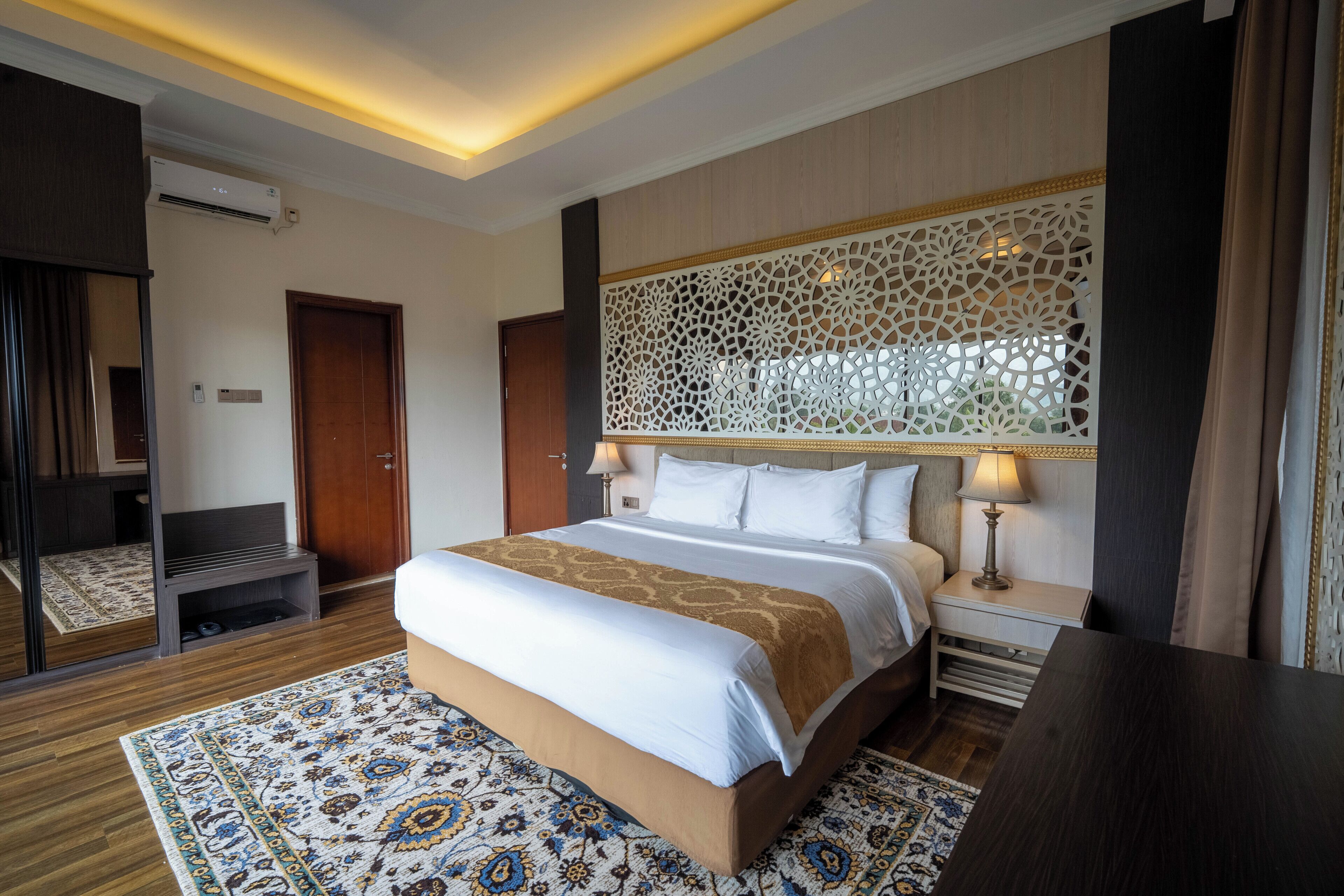 A hotel room with a large bed, a decorative headboard, a nightstand with a lamp, a wooden floor, and a patterned rug.