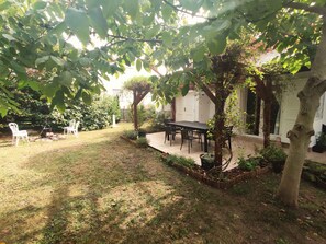 Un patio con mesa y sillas, rodeado por un jardín con diversas plantas y árboles.