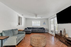 A living room with a grey sofa, a wicker ottoman, a wooden side table, and a TV mounted on the wall.