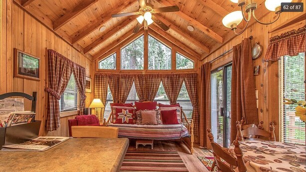 Un acogedor interior de cabaña con techo de madera, una cama con ropa de cama a rayas, una mesa de comedor y un ventanal con vistas al bosque.