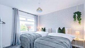Ein Hotelzimmer mit zwei Betten, einem Fenster mit Vorhängen, einer hängenden Pflanze, einer Nachttischlampe und einem kleinen Tisch.