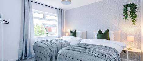 A hotel room with two beds, a window with curtains, a hanging plant, a bedside lamp, and a small table.