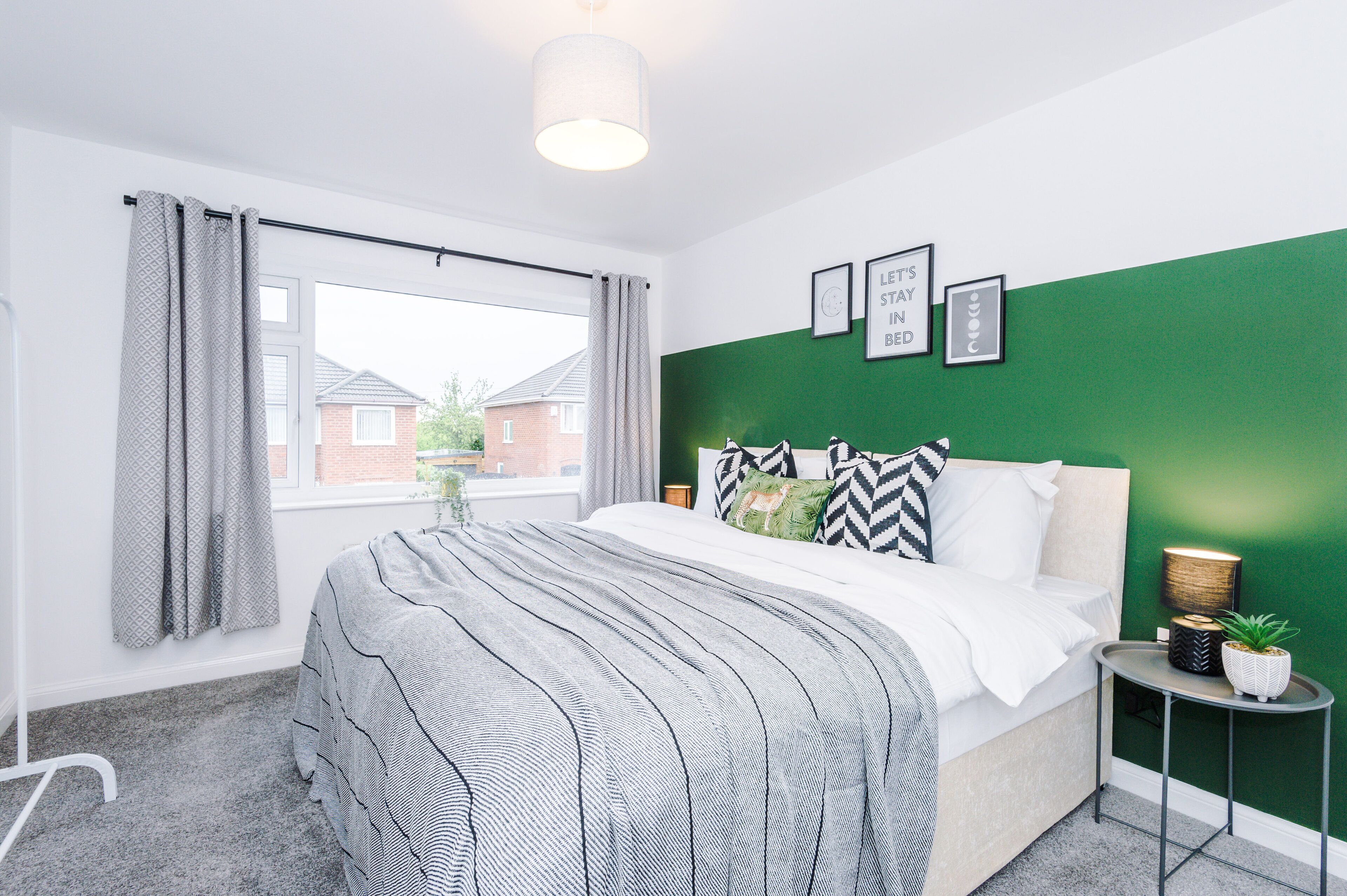 A bedroom with a bed, a window with curtains, a green accent wall, and a small bedside table with a lamp and a plant.