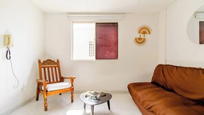 Un salon avec un fauteuil en bois, un canapé marron, une petite table sur laquelle se trouve un bol, et une fenêtre ornée d’un rideau rouge.