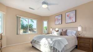 A bedroom with a large bed, two windows, a ceiling fan, and artwork on the walls.