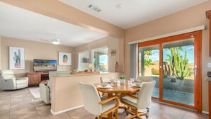 A spacious living area with a dining table, comfortable seating, a TV, and a view of a garden through a sliding glass door.