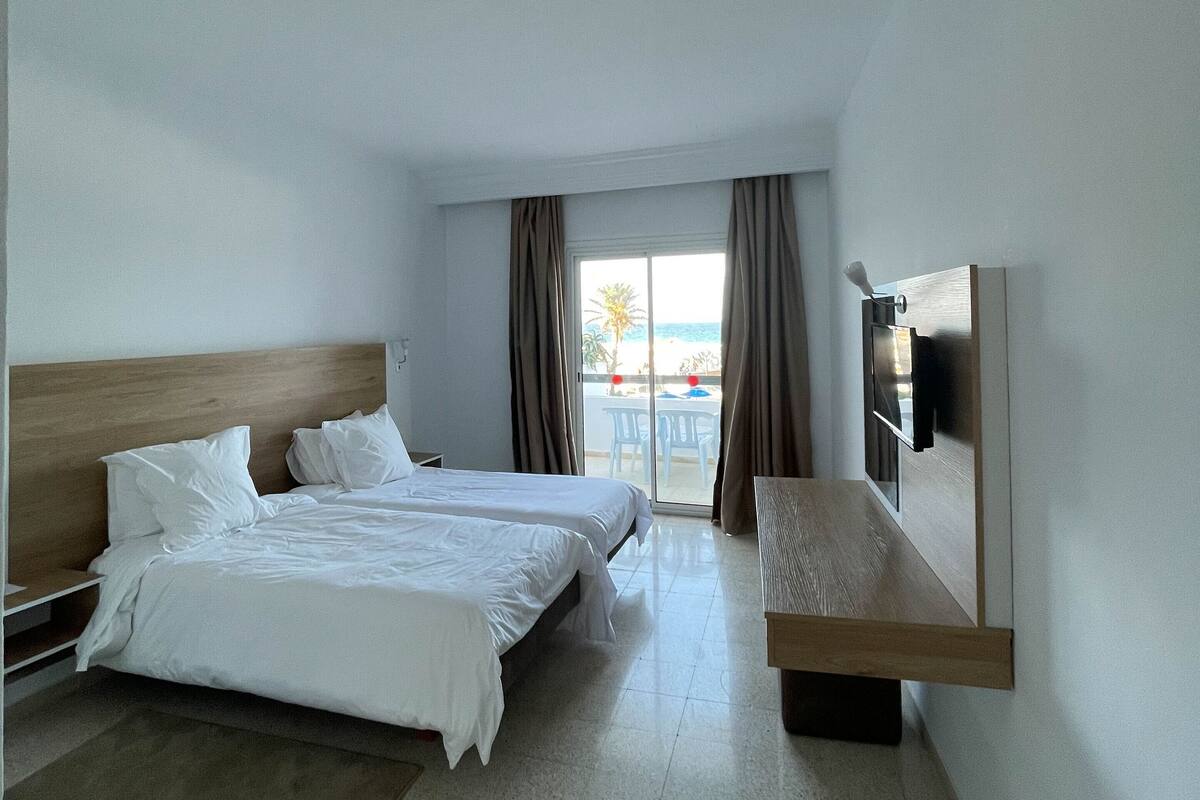 A hotel room with two beds, a TV, a wooden bench, and a view of the beach.
