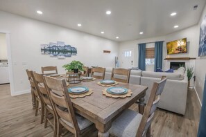 Un coin repas avec une table en bois dressée pour quatre personnes, entourée de chaises en bois. Une plante en pot est posée sur la table, et la pièce présente un design moderne avec un canapé et une cheminée en arrière-plan.